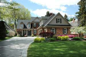 Spacious suburban house with a brick facade, gray roof, and manicured front lawn. A curved driveway leads to the entrance, surrounded by vibrant shrubs.